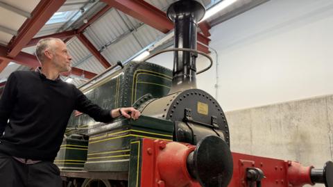 A man with grey hair wearing a black jumper and black trousers stands on the left next to a dark green and red locomotive engine. He is resting his elbow on the front of the train and looking up at it.