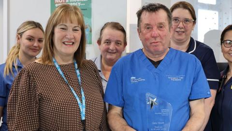 Gary Hewitt with short black hair and wearing blue hospital scrubs holds and engraved glass award next to him is trust chief executive Lyn Simpson who has shoulder length brown hair and a brown top and behind him stand a number of medical staff