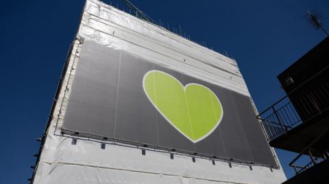 A tall tower block wrapped in white protective sheeting and scaffolding, with a large green heart banner mounted on one side, against a clear blue sky.
