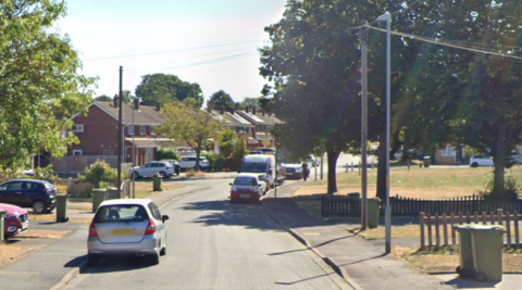 A residential street on a sunny day. Terraced houses are on one side of the street while there is a grassy area on the other. Cars and telegraph poles line both sides of the streets and residents have their bins out.