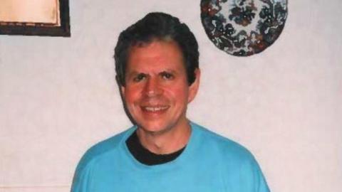 Stephen Ridgeway wearing a black t-shirt underneath a blue jumper. He has short black curly hair and is smiling at the camera. He is standing in front of a white wall which has a decorative plate and a picture frame displayed on it.