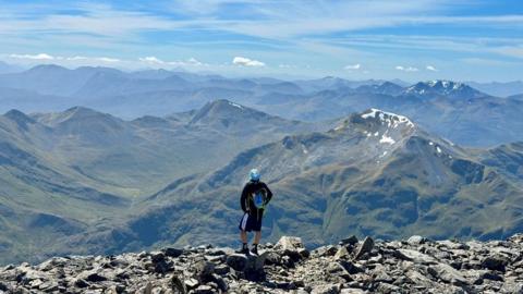 A man stands at the summit of Ben Nevis with a dramatic view of adjoining hills