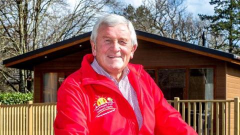 A man in a red fleece with short grey hair smiles at the camera. He is standing in front of a wooden chalet