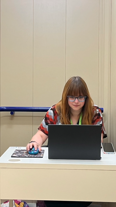 A woman with brown hair and a fringe sitting behind a laptop at a desk. She has set up her own personal office inside a lift.