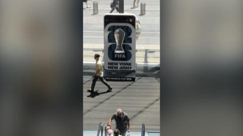 A man walking past what appears to be a face in a whole stand showing the FIFA world cup. It reads 'FIFA New York New Jersey'.