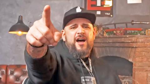 El Temach wears a black hoodie, a black and white cap and has a beard - he is pointing and shouting at the camera 