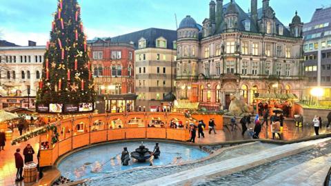 A wide scene of a city square with a fountain in the foreground, surrounded by wooden market stalls. A large Christmas tree sits in front of the surrounding city buildings.