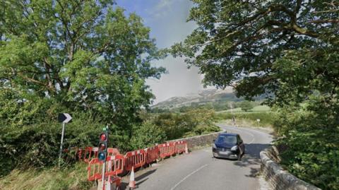The A683 over Straight Bridge near Sedburgh in Cumbria. It is a single carriageway road with low brick walls behind which are large trees. There is a temporary traffic light set to red, with a traffic cone and red temporary barriers covering gaps in the brickwork. A small black car is travelling in the middle of the road towards the camera. In the distance is a range of mountains.