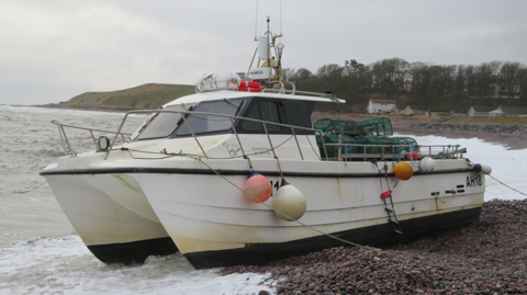 The Sea Eagle has a catamaran-like hull with a small cabin and there are green creels on the desk. The boat is on a shingle beach.