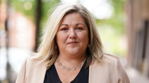 Fiona Elliott has short, blonde hair and has a neutral expression on her face as she walks outside the Clayton Hotel in Belfast. She is wearing a necklace with circular charms on it, as well as a beige suit jacket and a black v-neck top.