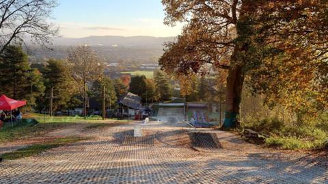 A view from the midway point of an artificial ski slope and ramp. There are trees to both sides and a landscape of hills, houses and a field in the background.