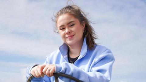 A woman wearing a blue zip up fleece sits on a vehicle. She is holding a steering wheel. Her hair is tied in a pony tail. A blue sky is behind her.