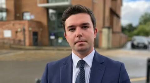 A man with short dark hair and a blue suit in front of a large blurred building