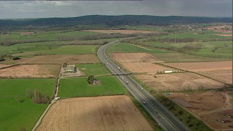 A drone shot of the proposed site of Marlcombe near Exeter Airport. It's a rural area with grass and farm land along with a busy road with two lanes in each direction. 