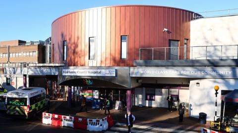 The main entrance to the Royal Sussex County Hospital. People can be seen walking through the main doors and an ambulance is parked outside. Above the from canopy area is a large bronze-coloured, cylinder shaped block
