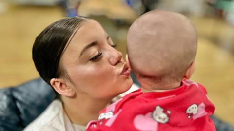 A woman is holding a baby, she has her hair tied back and is kissing the baby's cheek. The baby has the back of its head to the camera. The baby is wearing a red and pink outfit with hearts and kittens on.