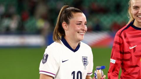 Ella Toone wearing England women's national football kit, which is white and blue. She is holding a drink on the pitch.