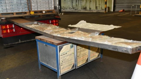 Bags of cannabis laid out inside a metal container on a table in a warehouse with large shutter doors in the background.