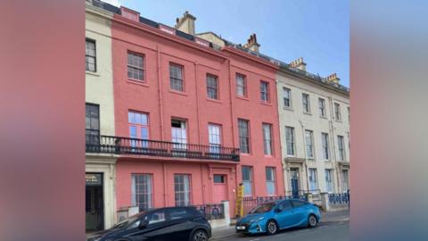 A row of terraced buildings on a street, with one property standing out prominently because it is painted a bold pink colour. The pink building has three storeys plus an additional level set back on the roof, and its windows and door frames are also painted in matching shades of pink. A black metal balcony runs across part of the first floor, and decorative architectural details are visible on the façade.
The neighbouring buildings on either side are painted in pale cream or off‑white. The street in front of the houses has a couple of parked cars, including a dark‑coloured car and a bright blue one. The sky above is clear and blue.