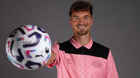 Thomas Meunier holds a ball outstretched in his right hand. He is wearing a pink Sunderland jersey