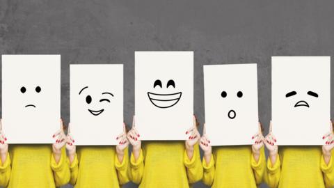 Five women wearing bright yellow jumpers standing in a row each holding a large rectangle white piece of paper over their face expressing a different emotion