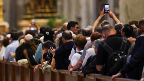 Queue of people standing against short barrier, seen from behind. One person raises their phone up in the air to take a picture of the crowd