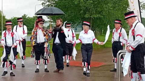 A group performs a traditional morris dance in costume. The seven men and one woman are wearing dark trousers, black hats with red and white ribbons, white shirts and are twirling white cloths.