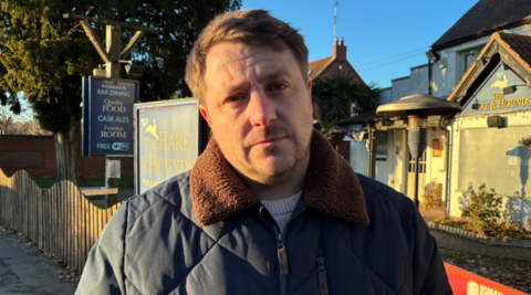 A man in a dark coat is looking at the camera. He is in front of a pub site, which has the name The Hare & Sounds on a board and on the outside of the building.