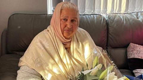 A old woman with grey hair peeking out of a pale pink head scarf. She has an ivory scarf with white embroidery wrapped across her shoulders. she is sat on a chocolate brown leather sofa while holding a bouquet of white lilies