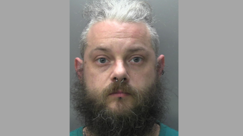 A mugshot of Jamie Elliott following his arrest. Jamie has greying hair, a big beard and is wearing a nose ring. 