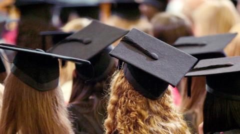 Students sitting with black graduation hats on their heads. The picture is taken from behind them. They have long hair which is blonde, brown or black. 