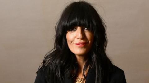 Claudia Winkleman wearing black clothes and gold necklace looking at camera.
