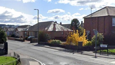 A town street with a sign in the foreground and two brown brick houses with trees cars in the driveways and a blue cloudy sky on a sunny day