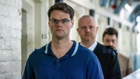 A man in a blue shirt wearing black rimmed glasses with a prison guard behind him