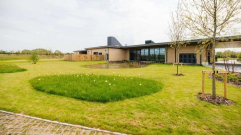 One storey building made of beige brick with large dark windows. There is grass in front with wildflowers, two trees and a path. There is also a pond and then trees in the distance.