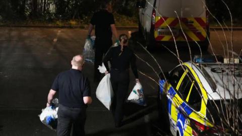 Three people walking with bags towards a police car.