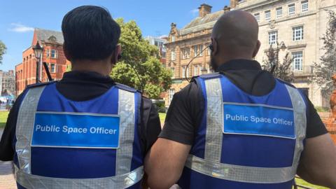 The backs of two of the new officers in blue uniforms bearing the words 'Public Space Officer'