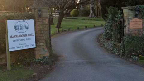 Sign which reads Bearwardcote Hall Residential Home to the left of a gate, with a road leading beyond the gates