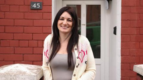 Lisa Molloy, 42, inside the doorway of her home on Trinity street. She has black long straight hair, brown eyes, and smiles with her teeth at the camera. She wears a light brown vest top and a cream cardigan with pink love hearts.