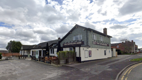 The pub stands back from a road with a large car park on the left of the image.It has greenery decor by its entrance and a small outdoor seating area. The top part of the pub is painted pale green with a cream lower half.