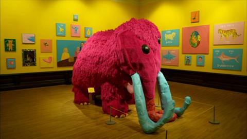 A life-size sculpture of a woolly mammoth, with pink fur, blue horns and yellow toenails in the center of the room. Colourful paintings hang on the yellow walls of an art gallery, with wooden floor.