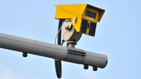 A yellow speed camera attached to a grey pole in the air.