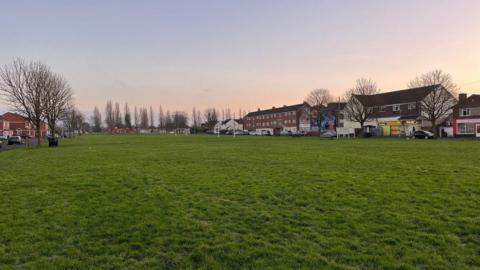 A large public green space surrounded by houses, flats and some shops and businesses in a deprived area of Gloucester. The sky is a wintry sunset and the trees are bare.