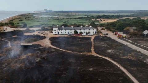 A lone two-storey house surrounded by fields, the ones in front entirely scorched by wildfires. To the left of the frame are sandbanks and the sea
