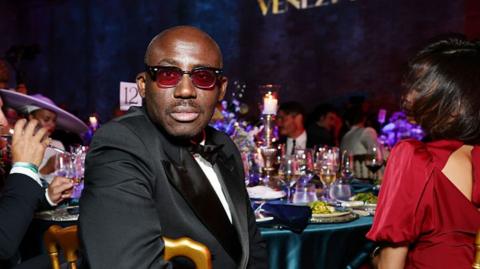 Edward Enninful looks back at the camera from a dinner party table, a women in red sits next to him. 