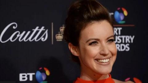 Woman with brown hair with her hair up smiling and wearing red clothing. There is a dark backdrop behind from what appears to be an earlier awards event.