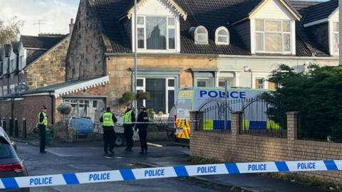 Police officers behind police tape are standing next to a police van outside a house