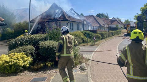 Two fire officers wearing protective pale brown overalls with hi-visibility markings in acid yellow are in a residential street lined with several bungalows. Well-maintained hedging lines the front of the properties. One of the roofs is gutted with gaping holes and billowing smoke. A fire engine with a hydraulic ladder is further up the road and a crew member is pointing a hose full of water at the burnt-out roof.  