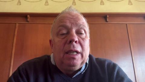 John Stiles is seen on a video link, talking to a reporter. He is middle-aged, has short light-coloured hair, and is wearing a shirt and jumper