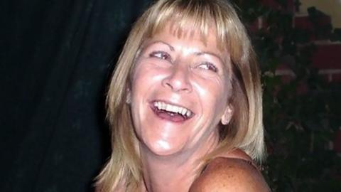 Jean Hanlon looking slightly off camera. She has shoulder-length blonde hair and is laughing. Her top row of teeth are visible. She has light-coloured eyes. Only her shoulders, neck and head can be seen.